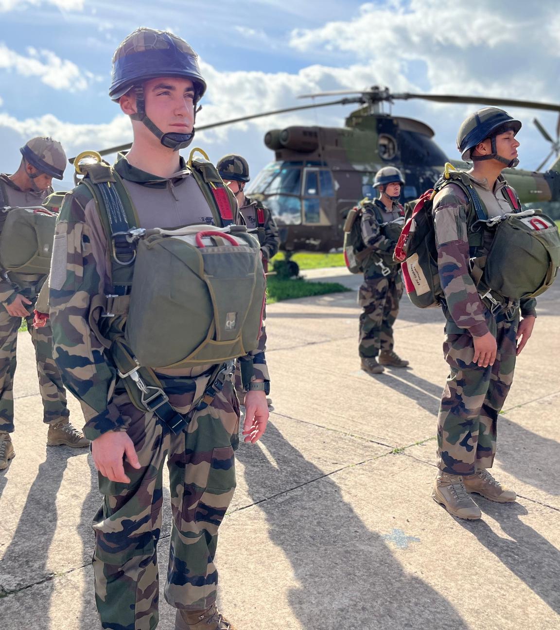 Mehrere Legionäre stehen vollausgerüstet auf einem Platz und warten auf den Materialcheck. Im Hintergrund steht ein Helikopter.