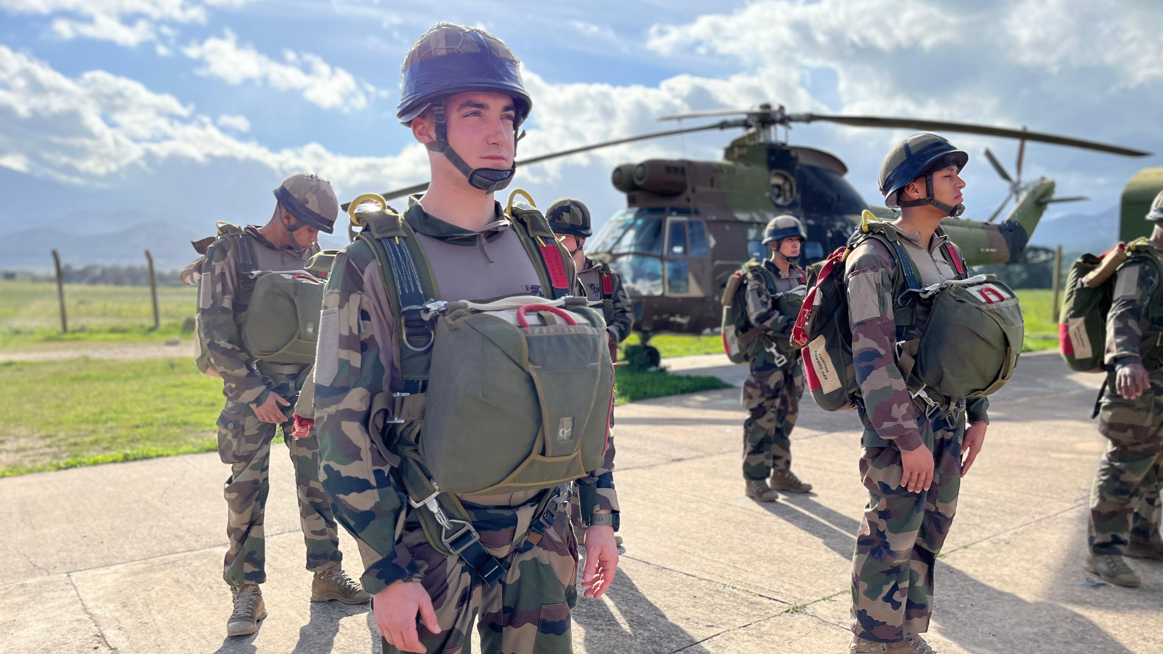 Mehrere Legionäre stehen vollausgerüstet auf einem Platz und warten auf den Materialcheck. Im Hintergrund steht ein Helikopter.
