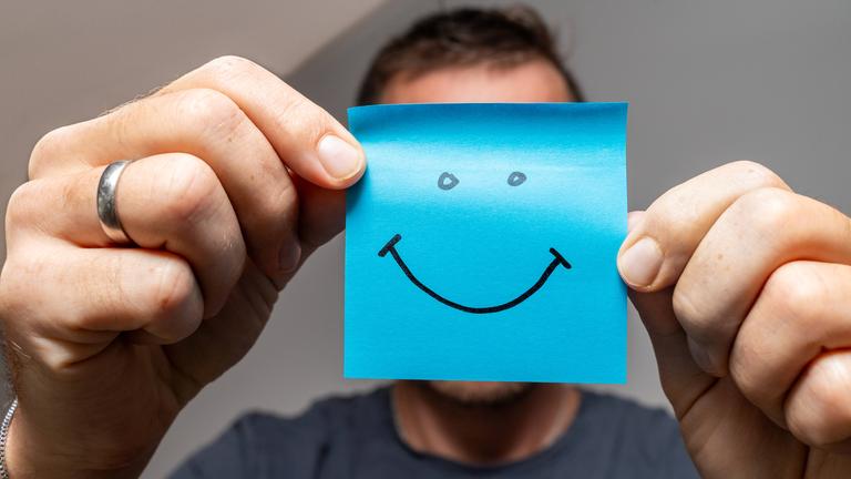 Ein Mann hält eine blaue Haftnotiz, auf die ein Smiley gemat ist, vor sein Gesicht.