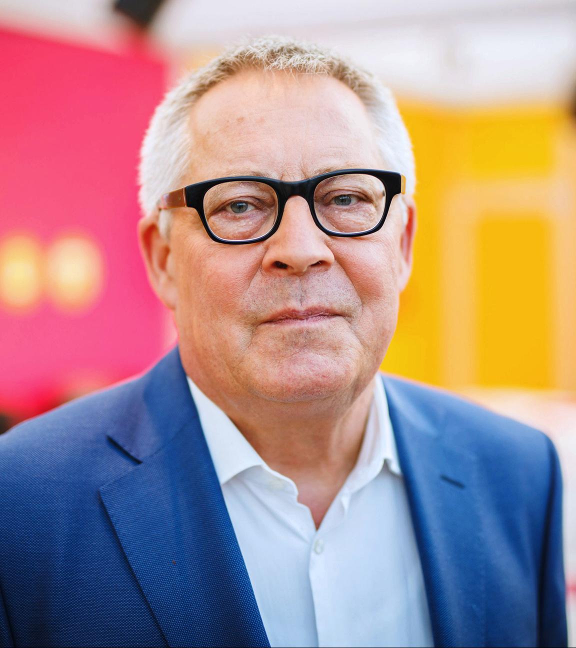 Porträt Karl Schlögel: Der Historiker blickt in die Kamera, trägt eine Brille und einen dunklen Blazer. Der Hintergrund ist neutral in den Farben orange und rot.