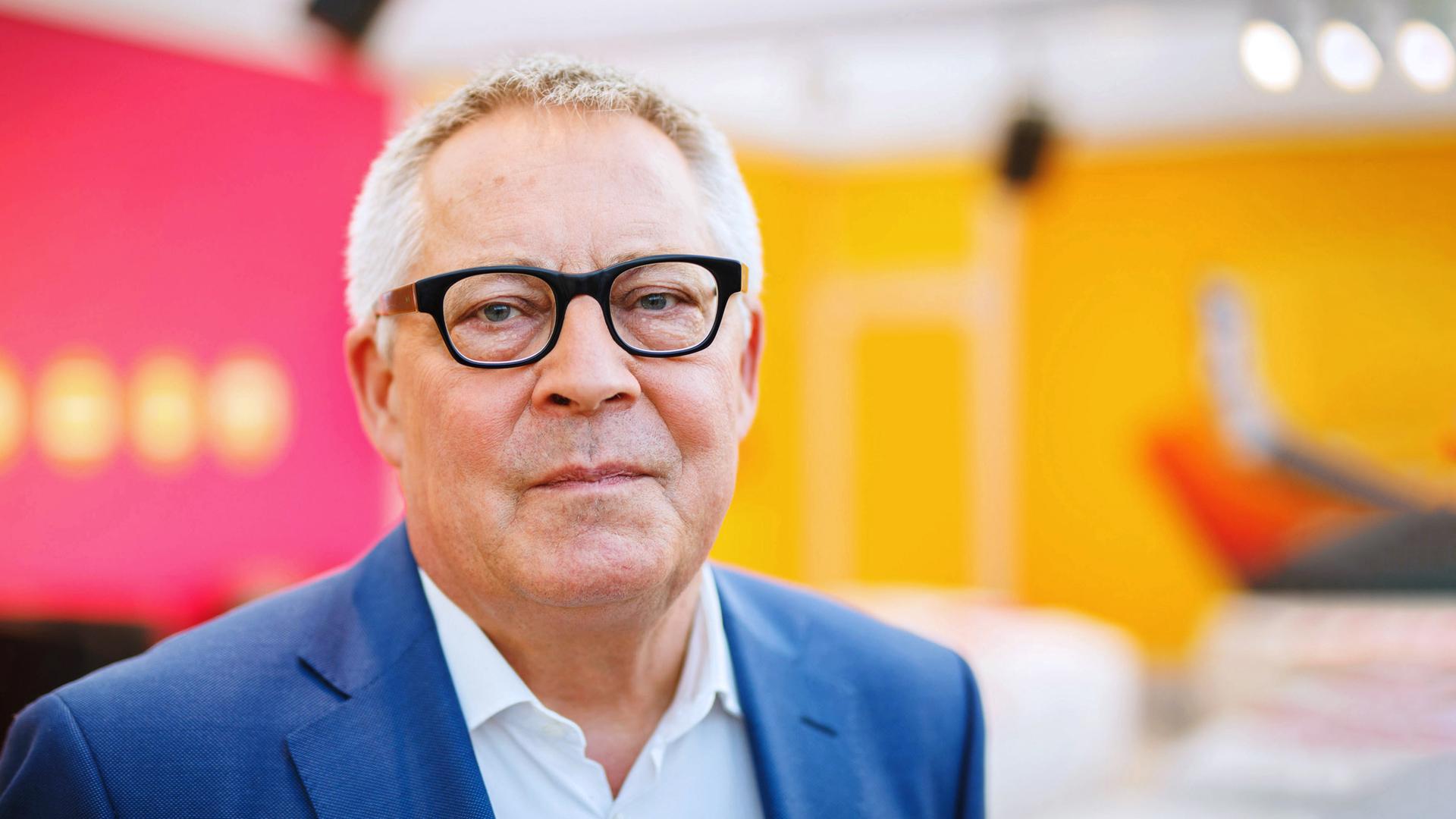 Porträt Karl Schlögel: Der Historiker blickt in die Kamera, trägt eine Brille und einen dunklen Blazer. Der Hintergrund ist neutral in den Farben orange und rot.