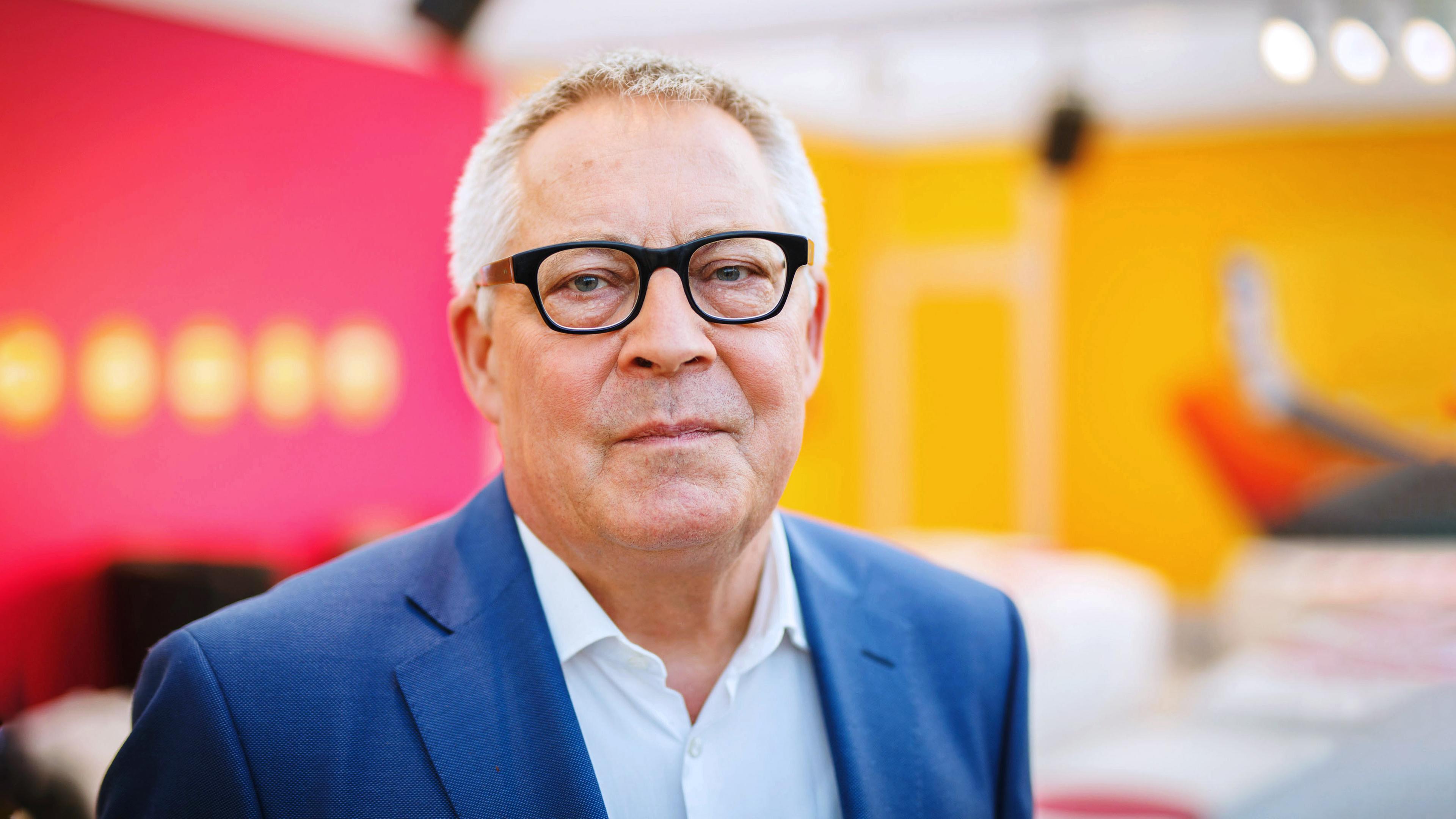 Porträt Karl Schlögel: Der Historiker blickt in die Kamera, trägt eine Brille und einen dunklen Blazer. Der Hintergrund ist neutral in den Farben orange und rot.