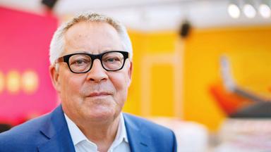 Porträt Karl Schlögel: Der Historiker blickt in die Kamera, trägt eine Brille und einen dunklen Blazer. Der Hintergrund ist neutral in den Farben orange und rot.