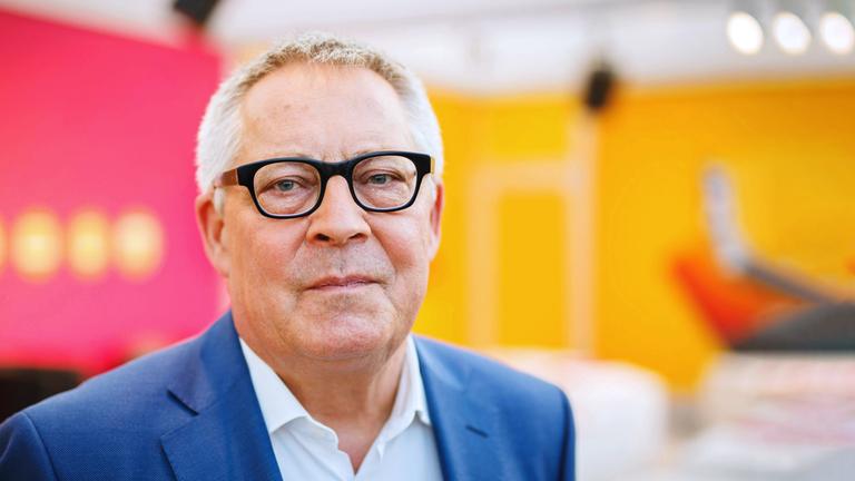 Porträt Karl Schlögel: Der Historiker blickt in die Kamera, trägt eine Brille und einen dunklen Blazer. Der Hintergrund ist neutral in den Farben orange und rot.