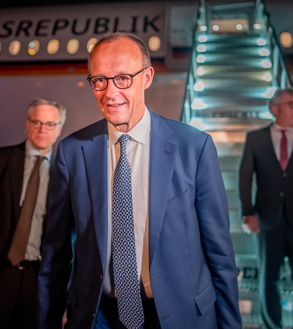 Mehrere Männer in Anzügen stehen in der Dämmerung vor einem Flugzeug, an das eine Rolltreppe angedockt ist.