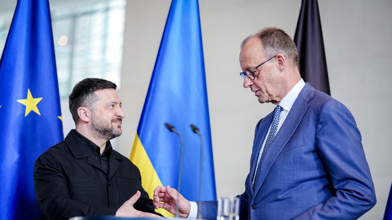 Bundeskanzler Friedrich Merz und Wolodymyr Selenskyj, Präsident der Ukraine, geben im Bundeskanzleramt bei den deutsch-ukrainischen Regierungskonsultationen eine Pressekonferenz. Sie stehen vor einem Mikrofon-Pult und sind im Begriff sich die Hände zu geben.