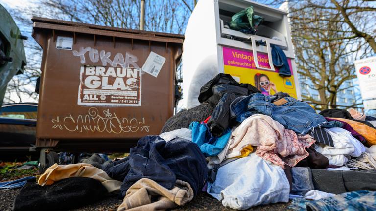 Ein überfüllter Altkleider-Container. Vor dem Container liegen bereits einige Müllsäcke mit Kleidung, die nicht mehr in den Container gepasst haben.