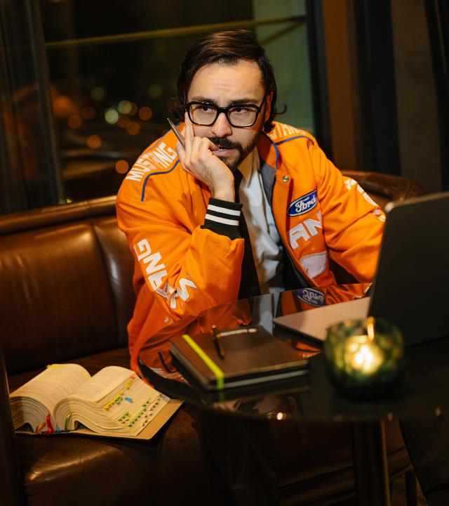 Furkan sitzt auf einem Sessel und er schaut grübelnd zur Seite. Vor ihm steht sein Laptop auf einem Tisch und ein "dicker Wälzer" liegt daneben. 