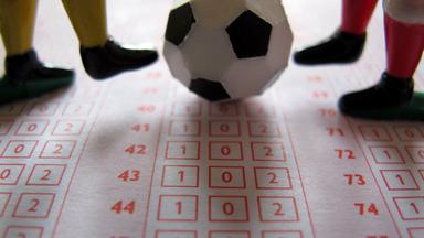 Die Füße von zwei Spielfiguren spielen mit einem kleinen Fußball. Der Untergrund ist das Blatt eines Tippspiels.