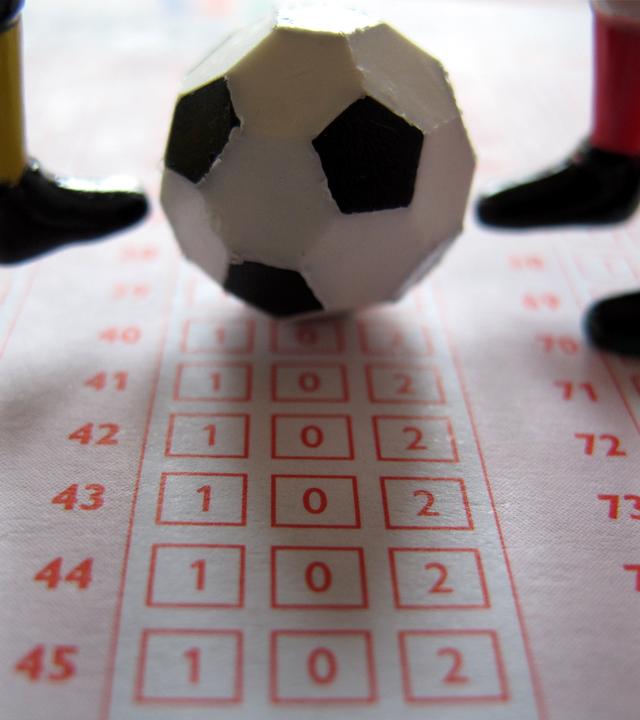 Die Füße von zwei Spielfiguren spielen mit einem kleinen Fußball. Der Untergrund ist das Blatt eines Tippspiels.
