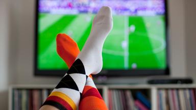 Überschlagene Beine vor einem Fernseher, der ein Fußballfeld zeigt