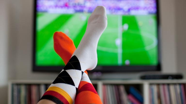 Überschlagene Beine vor einem Fernseher, der ein Fußballfeld zeigt