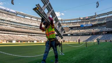Ein Arbeiter trägt auf dem Rasen des NYNJ-Stadion in den USA eine Leiter. Im Hintergrund sieht man weitere Vorbereitungen für die Fußball-Weltmeisterschaft 2026.
