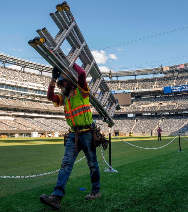 Ein Arbeiter trägt auf dem Rasen des NYNJ-Stadion in den USA eine Leiter. Im Hintergrund sieht man weitere Vorbereitungen für die Fußball-Weltmeisterschaft 2026.