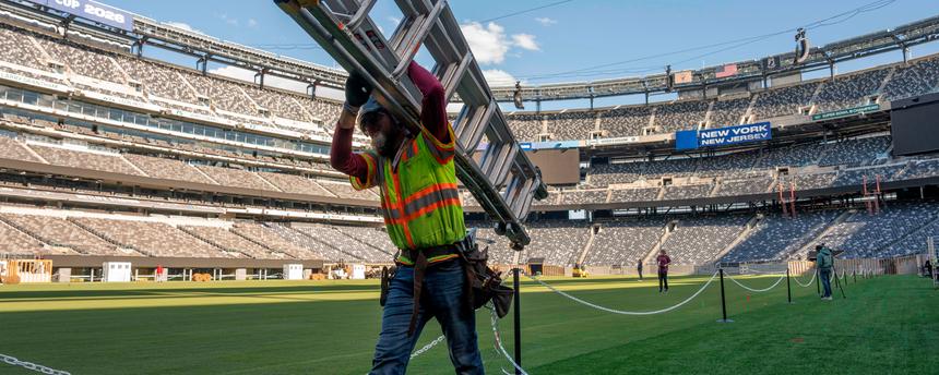 Ein Arbeiter trägt auf dem Rasen des NYNJ-Stadion in den USA eine Leiter. Im Hintergrund sieht man weitere Vorbereitungen für die Fußball-Weltmeisterschaft 2026.