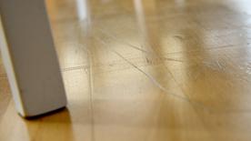 La pata de una silla blanca descansa sobre un suelo de parquet claro, a la derecha del cual se pueden ver profundos arañazos.