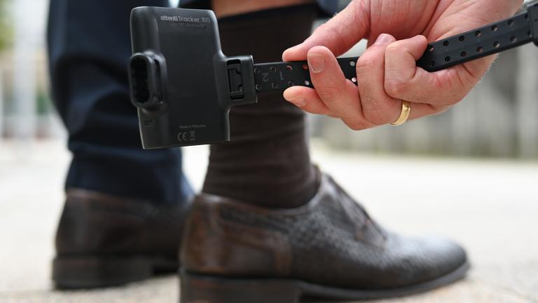 Ein Mann hält zu Demonstrationszwecken eine elektronische Aufenthaltsüberwachung, bekannt als elektronische Fußfessel, vor das Bein eines Probanden. 