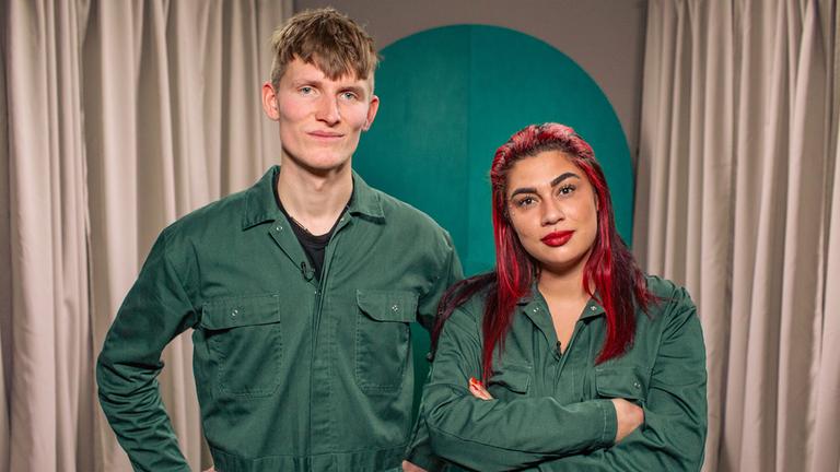Ein Mann und eine Frau in grünen Overalls stehen nebeneinander und blicken in die Kamera. Der Mann hat die Arme in die Hüfte gestemmt, die Frau hat rote Haare und die Arme verschränkt. Beide lächeln.