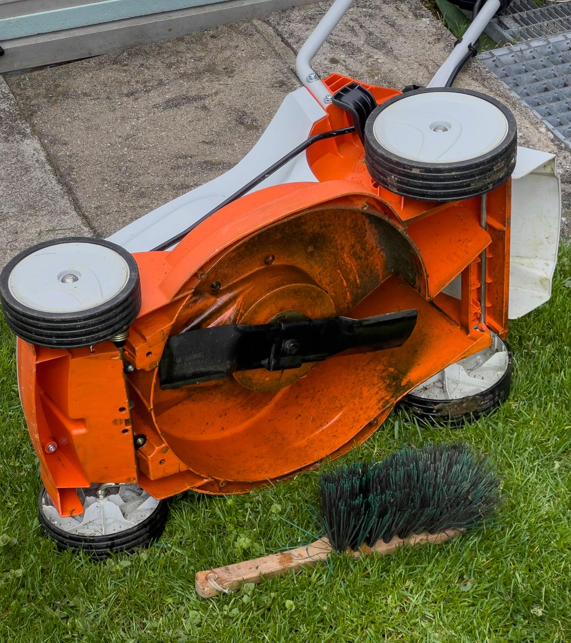 Ein Rasenmäher liegt auf der Seite, daneben liegt eine Reinigungsbürste uaf dem Boden.