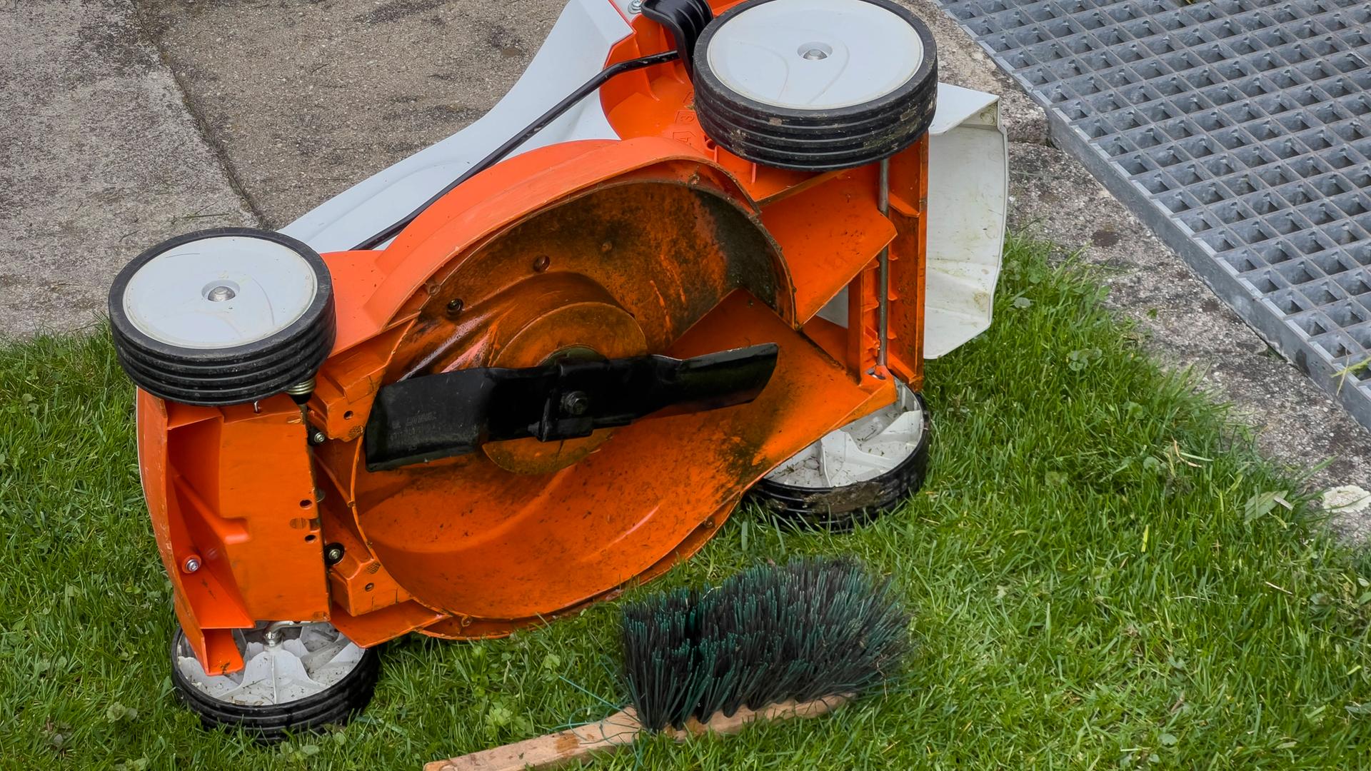 Ein Rasenmäher liegt auf der Seite, daneben liegt eine Reinigungsbürste uaf dem Boden.