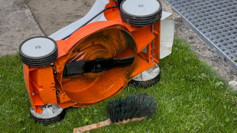 Ein Rasenmäher liegt auf der Seite, daneben liegt eine Reinigungsbürste uaf dem Boden.