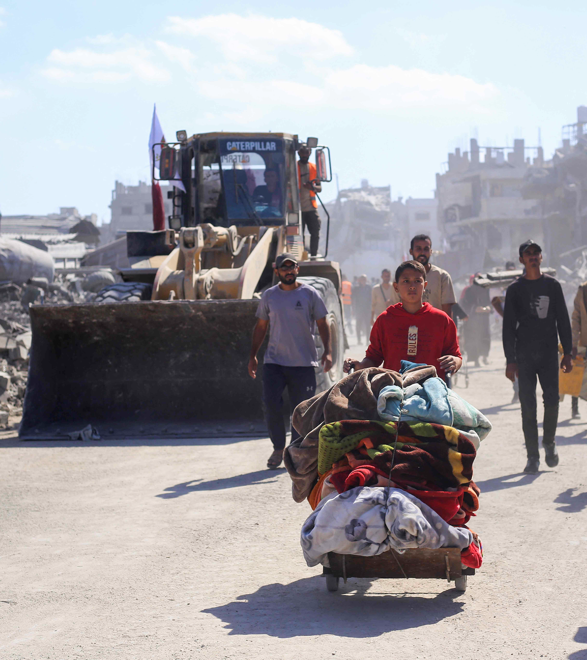 Menschen laufen durch eine zerstörte Stadt, Ein Junge schiebt einen Wagen mit seinen Habseligkeiten, am Straßenrand steht ein Bagger.