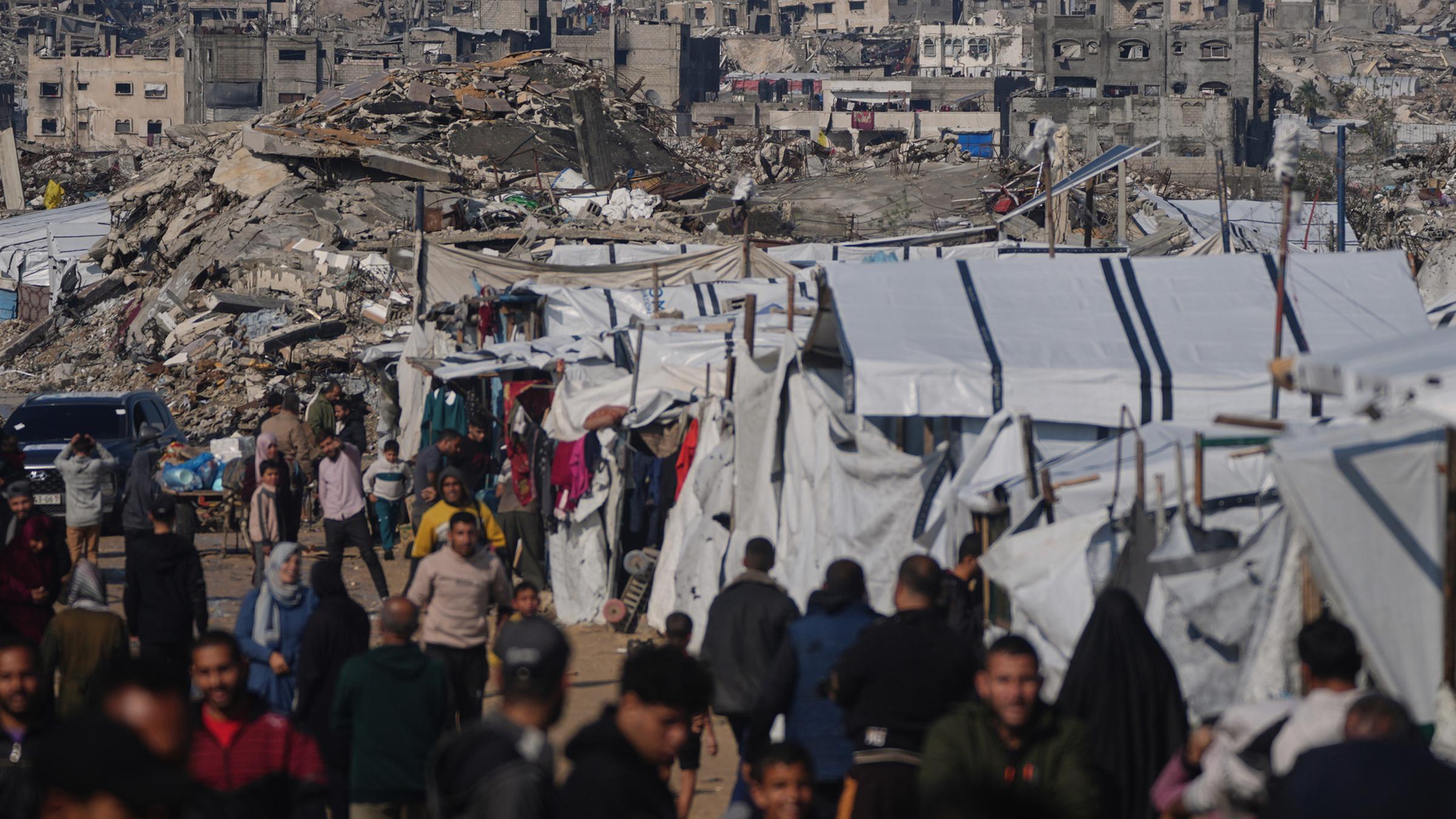 Palästinenser gehen an einem Zeltlager vorbei durch eine Straße in Gaza-Stadt, vor im Hintergrund zerstörten Gebäuden nach israelischen Luft- und Bodeneinsätzen, am 17. Dezember 2025.