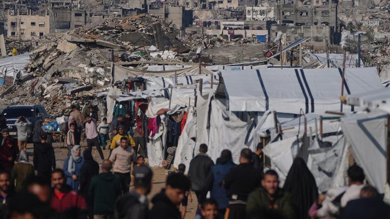 Palästinenser gehen an einem Zeltlager vorbei durch eine Straße in Gaza-Stadt, vor im Hintergrund zerstörten Gebäuden nach israelischen Luft- und Bodeneinsätzen, am 17. Dezember 2025.