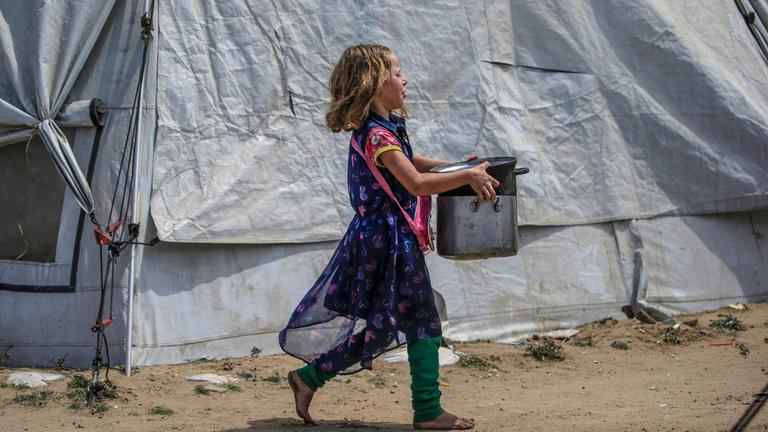 Ein intern vertriebenes palästinensisches Mädchen trägt Töpfe zu ihrer Familienunterkunft, nachdem sie Essen aus einer Wohltätigkeitsküche in Jabalia im nördlichen Gazastreifen erhalten hat.