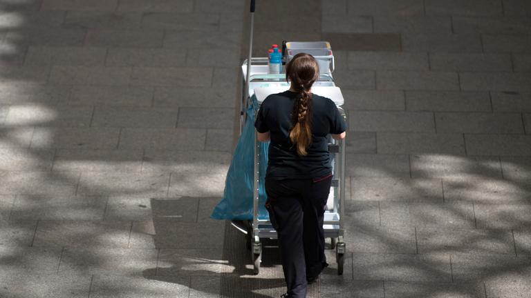 Symbolbild: Gebäudereinigerin schiebt Putzwagen vor sich her