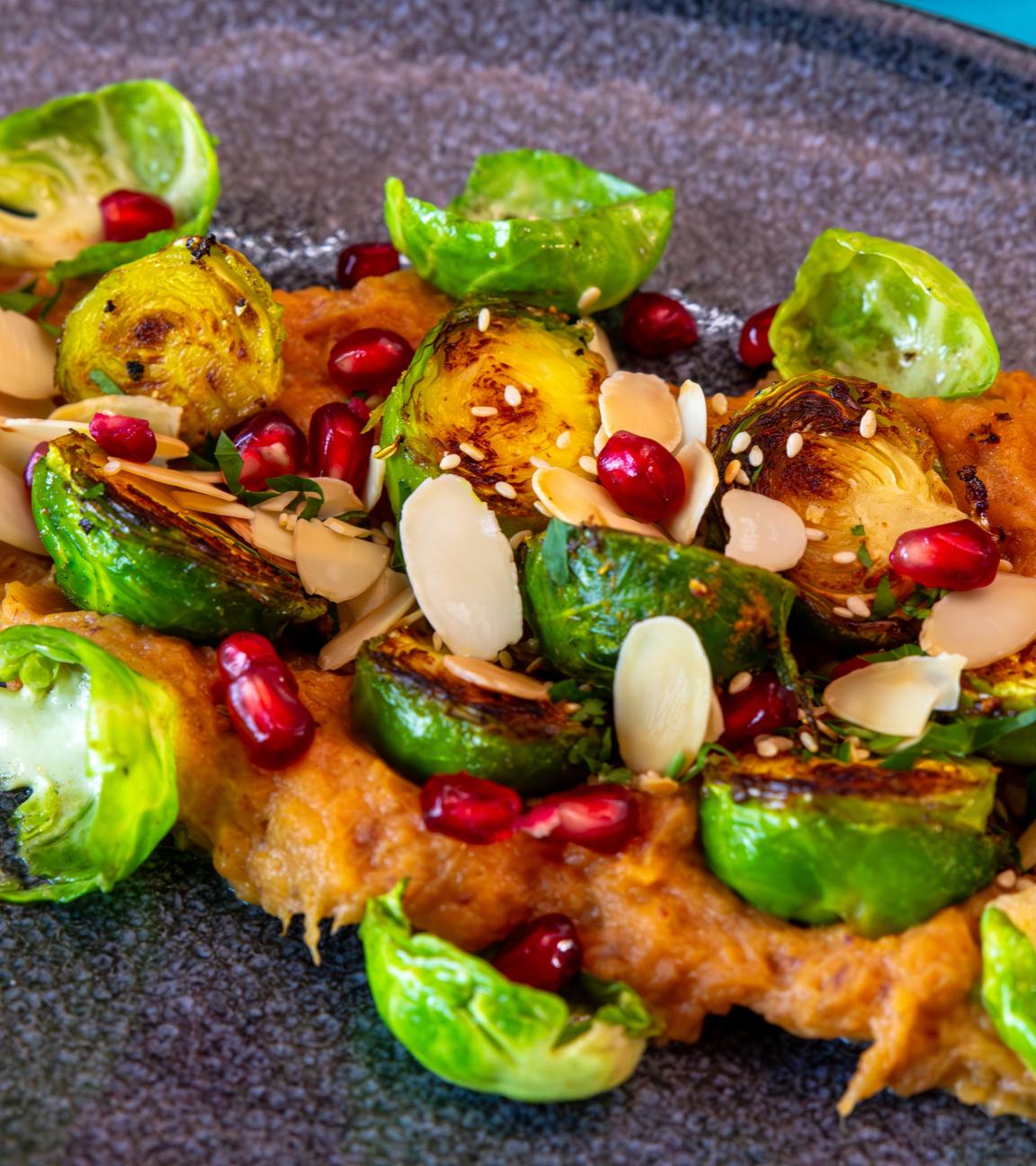 Teller mit gebratenem Rosenkohl auf Süßkartoffel-Dattel-Püree, getoppt mit knusprigen Mandelblättchen, Sesam und Granatapfelkernen.