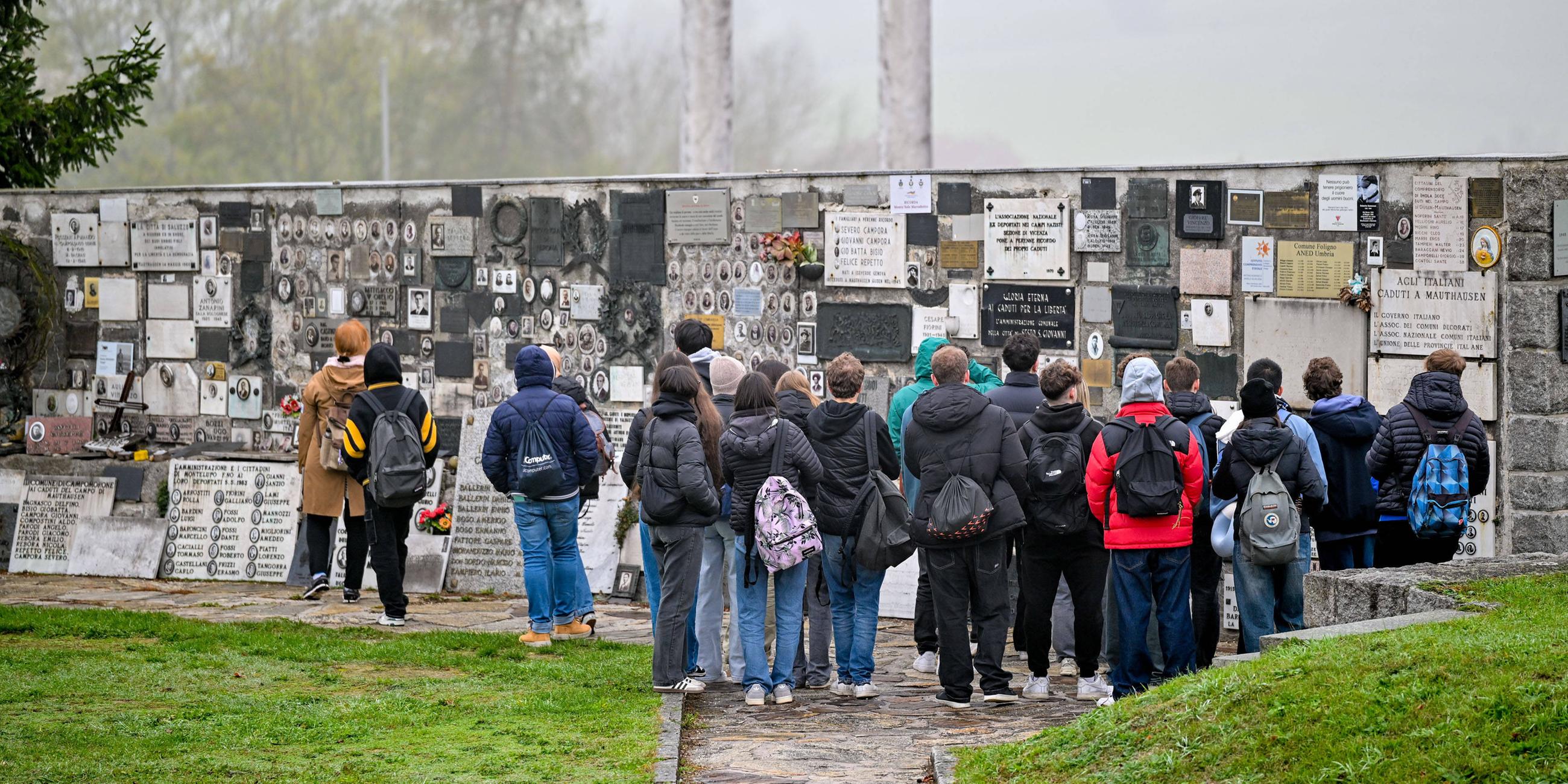 Schüler stehen im Freien vor einer Mauer, an der viele Tafeln angebracht sind.