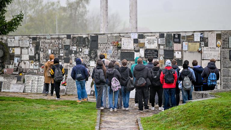 Schüler stehen im Freien vor einer Mauer, an der viele Tafeln angebracht sind.