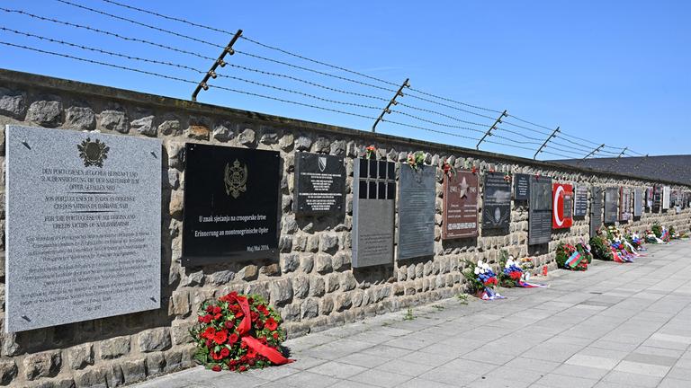 Eine Mauer mit einem Stacheldraht darüber. An der ganzen Mauer entlang hängen Gedenktafeln in verschiedenen Farben und Größen, alle sind rechteckig. Vor einigen Tafeln auf dem Boden liegen Blumenkränze.