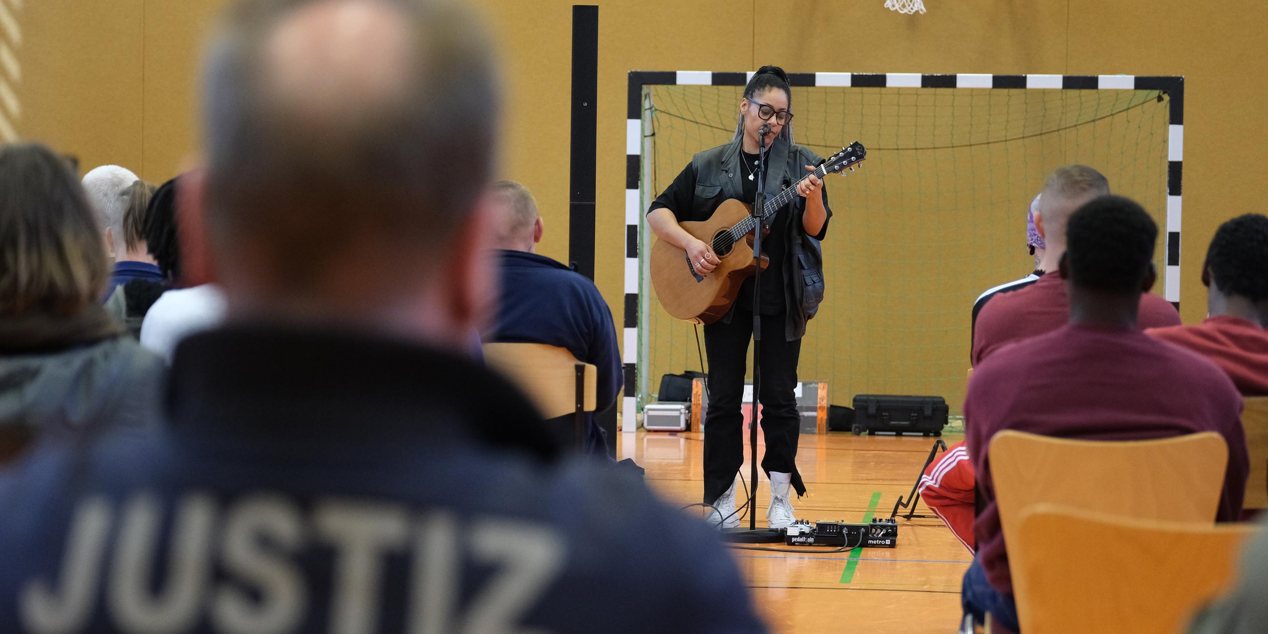 Eine Frau gibt mit einer Gitarre in der Hand ein Konzert in einer Turnhalle, mehrere Menschen sitzen auf Stühlen und schauen zu