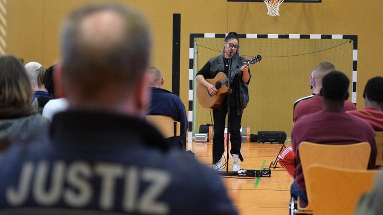 Eine Frau gibt mit einer Gitarre in der Hand ein Konzert in einer Turnhalle, mehrere Menschen sitzen auf Stühlen und schauen zu