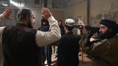 In einem heruntergekommenen Raum heben zwei Männer die Hände hoch, die von Taliban mit Waffen bedroht werden.