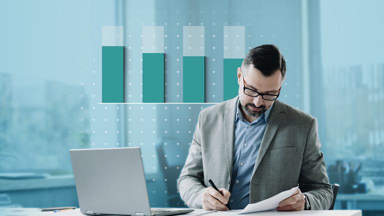 Ein Mann im Anzug sitzt an einem Schreibtisch vor einem Laptop. Im Hintegrund ist ein grafisches Balkendiagramm zu sehen.