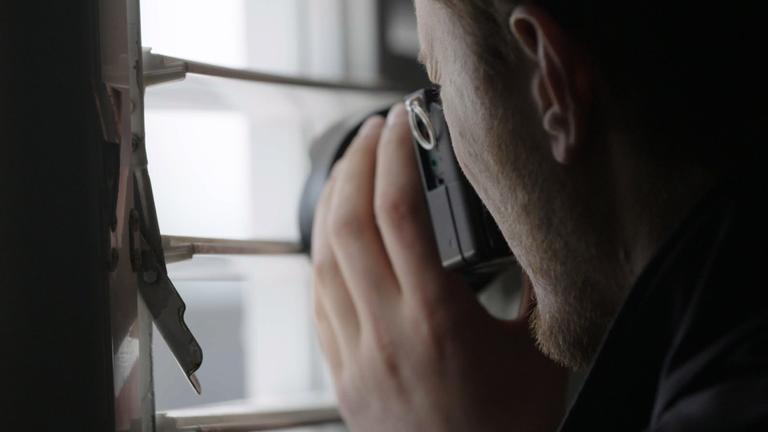 "Geheimes Israel - Der Mossad: Gefährliche Nachbarn": Ein Mann steht mit einer Kamera am Fenster und richtet das Objektiv durch die Lamellen einer Jalousie nach draußen (szenische Darstellung).