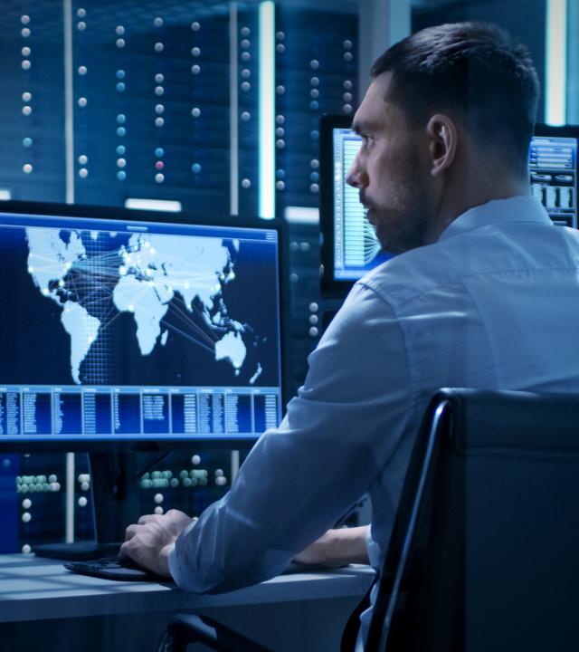 Ein Agent in einer moderner Einsatzzentrale, er sitzt am Rechner vor mehreren Monitoren, auf einem davon ist eine Weltkarte zu sehen.
