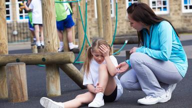 Ein Mädchen sitzt vor einem Klettergerüst auf dem Spielplatz und hält sich den Kopf. Eine Frau betreut sie.