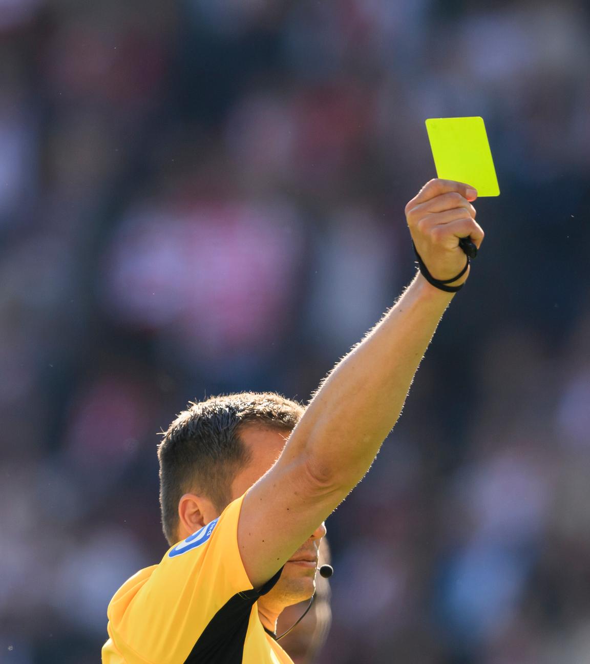 Ein Fußball-Schiedsrichter mit gelben Trikot reckt mit der rechten Hand eine gelbe Karte in die Höhe.