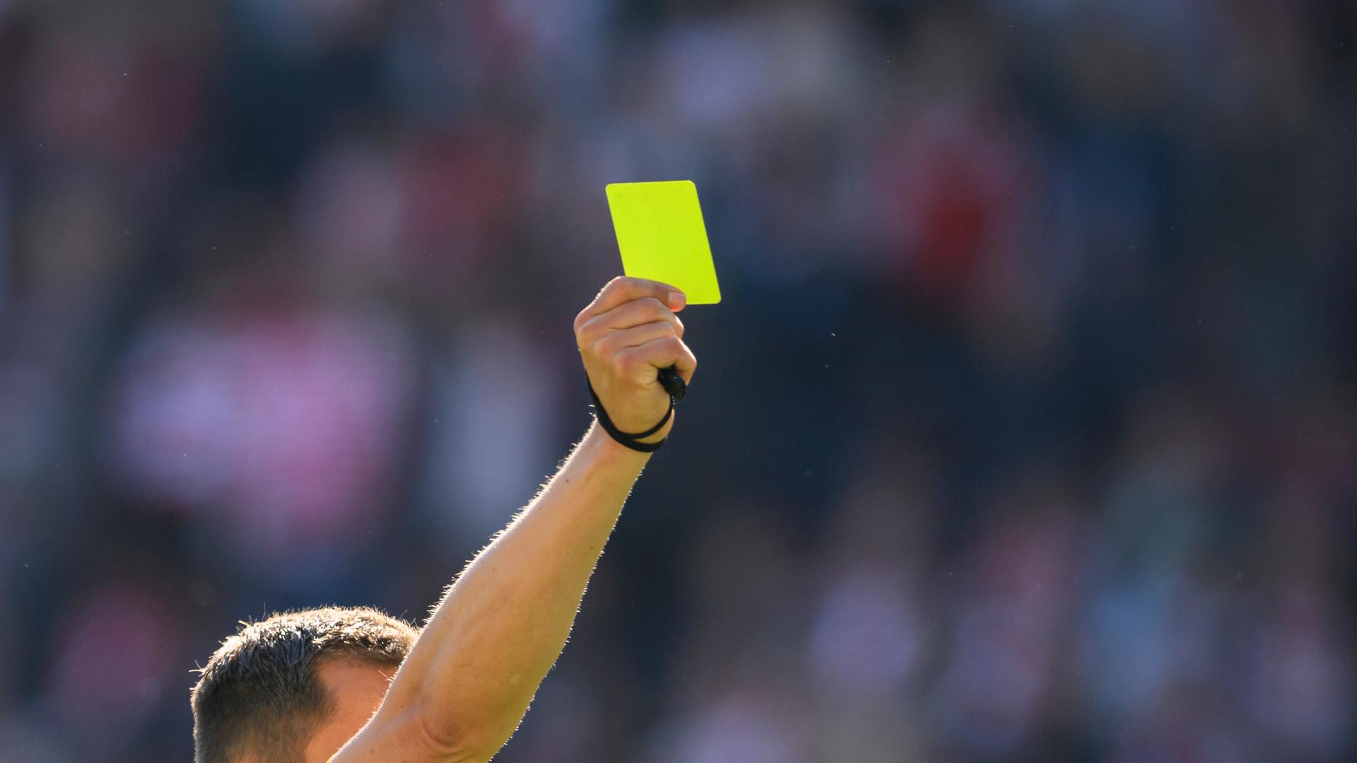 Ein Fußball-Schiedsrichter mit gelben Trikot reckt mit der rechten Hand eine gelbe Karte in die Höhe.