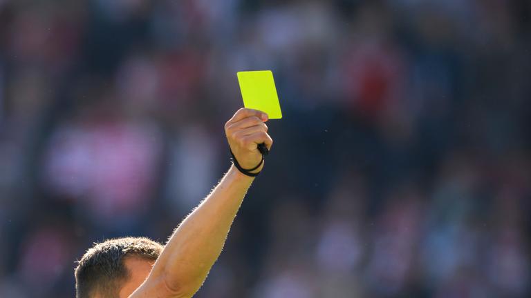 Ein Fußball-Schiedsrichter mit gelben Trikot reckt mit der rechten Hand eine gelbe Karte in die Höhe.