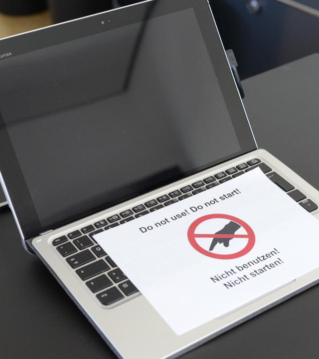 Auf einem Tisch befindet sich ein geöffneter Laptop mit schwarzem Bild. Darauf liegt ein Zettel mit dem Hinweis "Nicht benutzen!" und einer rot durchgestrichenen Hand.
