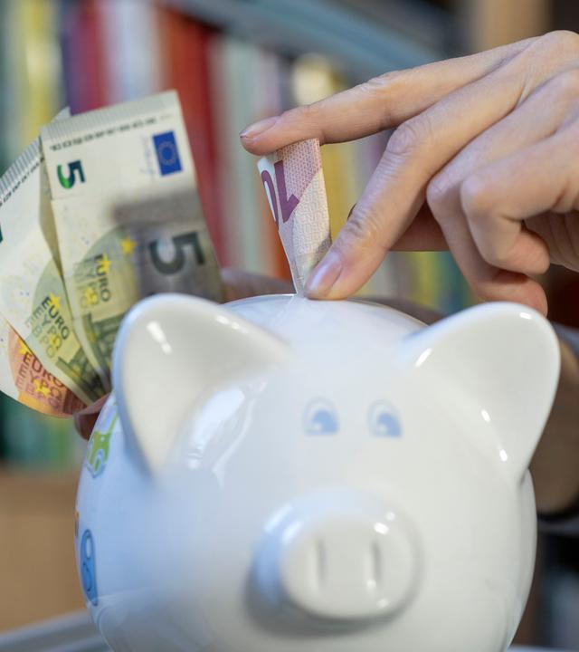 Eine Frau steckt einen Zehn-Euro-Schein in ein Sparschwein. Daneben sind weitere Geldscheine zu sehen.