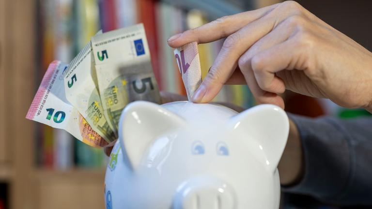 Eine Frau steckt einen Zehn-Euro-Schein in ein Sparschwein. Daneben sind weitere Geldscheine zu sehen.