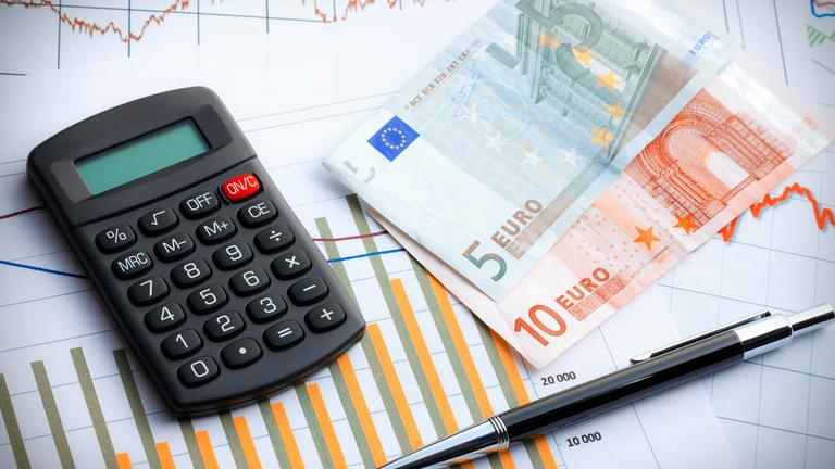 Geldscheine, ein Kugelschreiber und ein Taschenrechner liegen auf einem Papier mit einem ansteigenden Balkendiagramm.