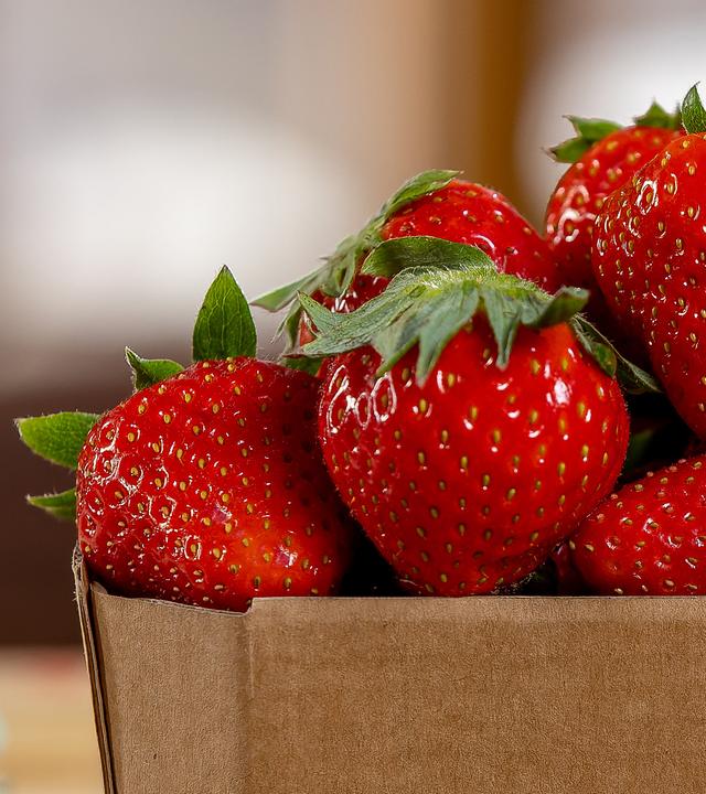 Im Anschnitt rechts eine braune Pappschale voll mit frischen reifen Erdbeeren.