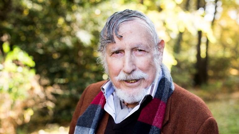 Georg Stefan Troller. Er trägt einen weißen Bart. Im Hintergrund sind Bäume und Büsche zu erahnen.